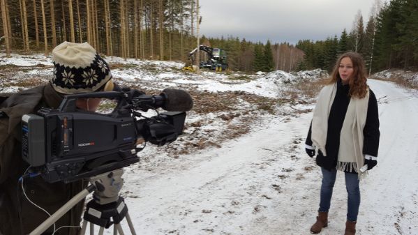 En person med stickad mössa filmer en tjej med en stor halsduk, i bakgrunden ses en avverkad skog samt en skogsmaskin