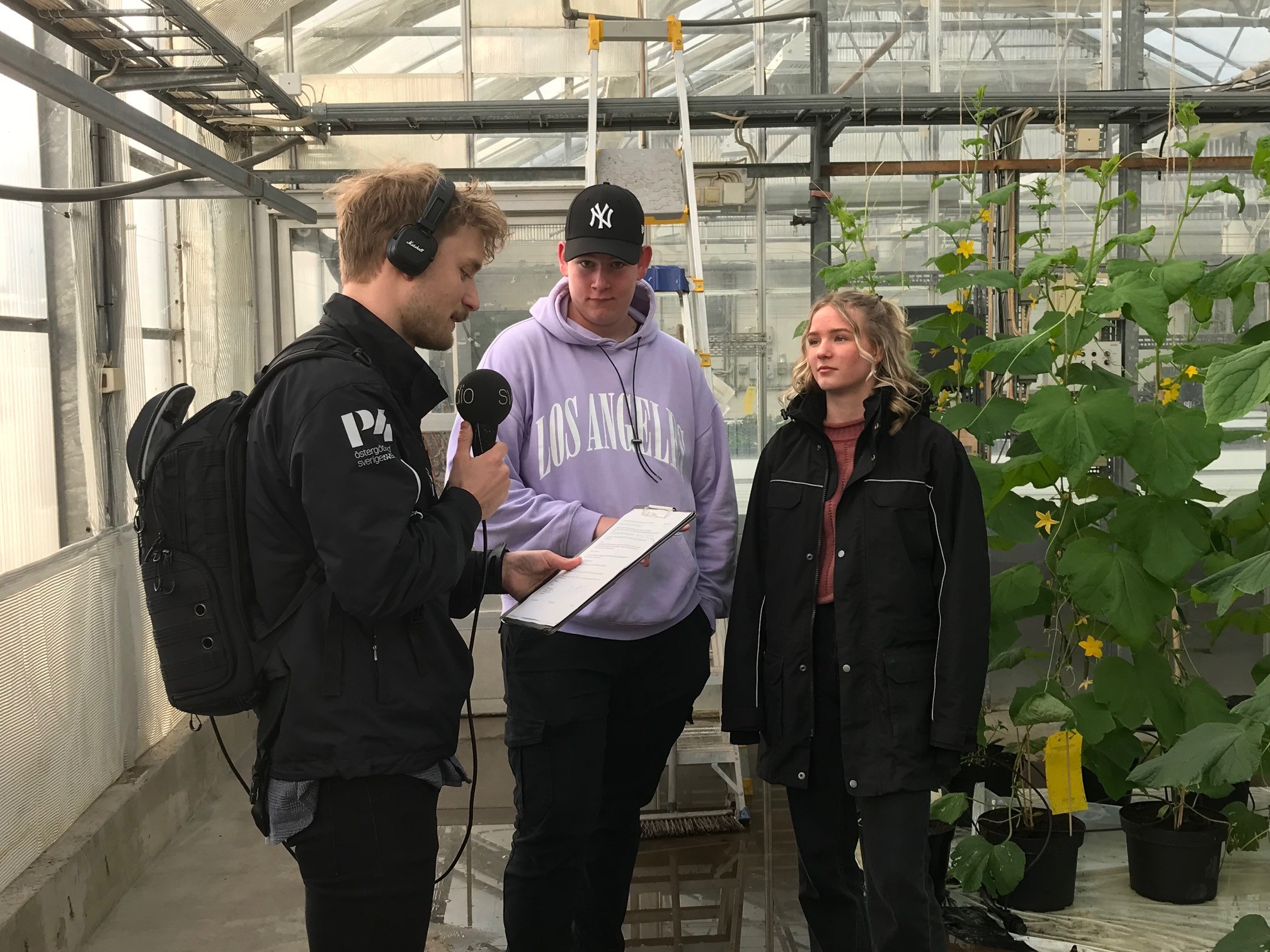 En radioreporter står i ett växthus och intervjuar en kille och en tjej
